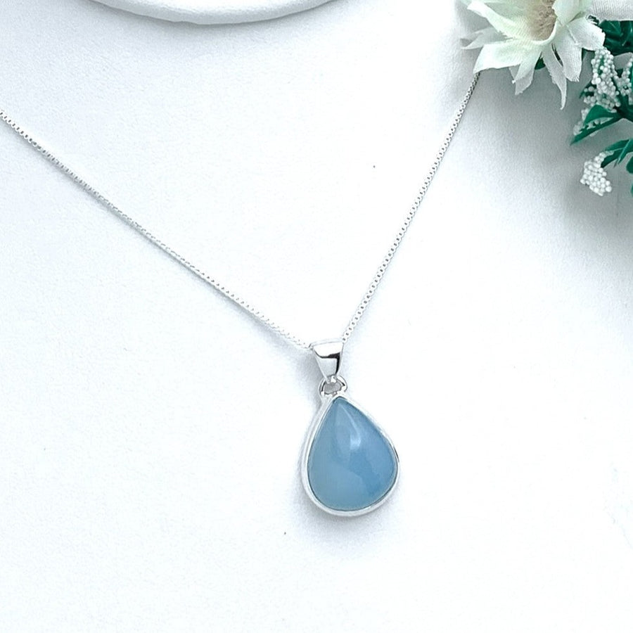 The Blue Chalcedony Pendant In A Perfect Silver Setting is a delicate sterling silver necklace featuring a teardrop-shaped pendant with a smooth, light blue semi-transparent Blue Chalcedony stone. The pendant hangs on an 18" box chain against a white background, adorned by a small arrangement of white flowers and green foliage.