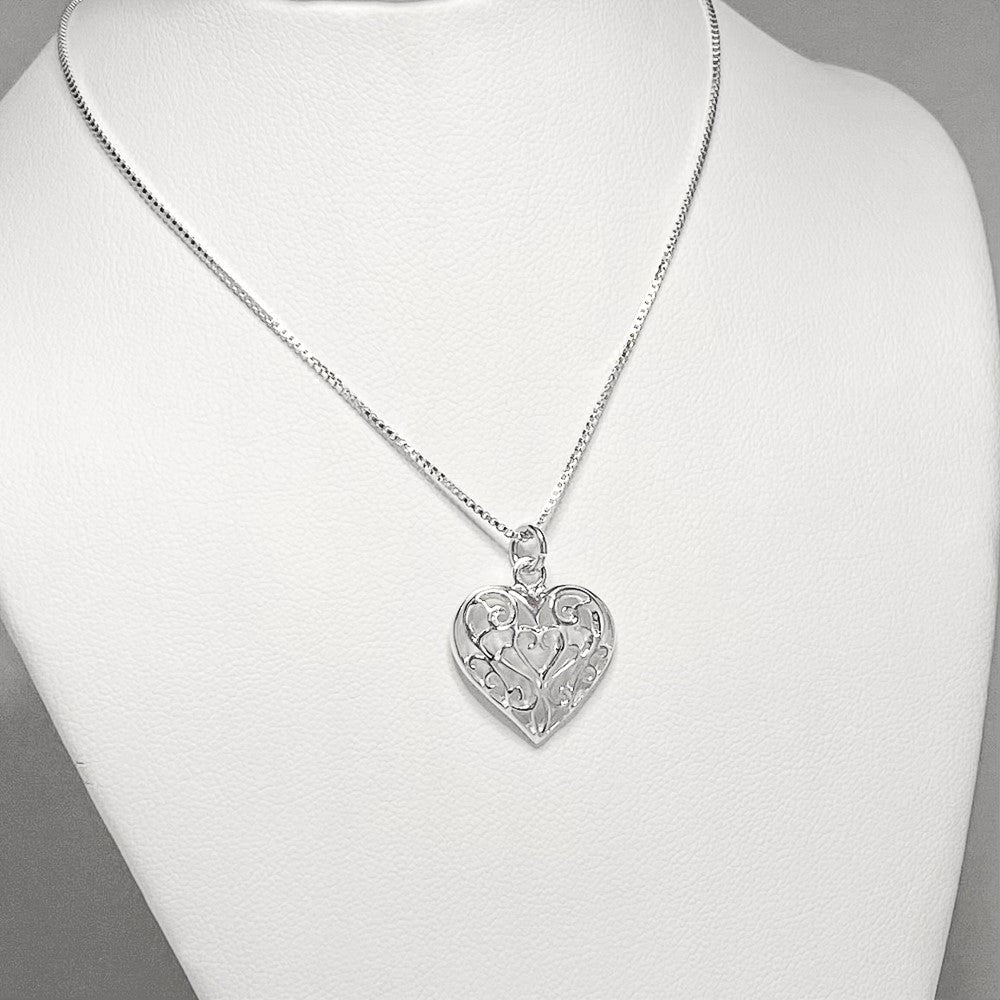 The Sterling Silver Filigree Heart Pendant, featuring intricate swirls and patterns, is elegantly displayed on a white mannequin bust. This exquisite piece includes an 18" sterling silver chain and comes with matching earrings to complete the set.