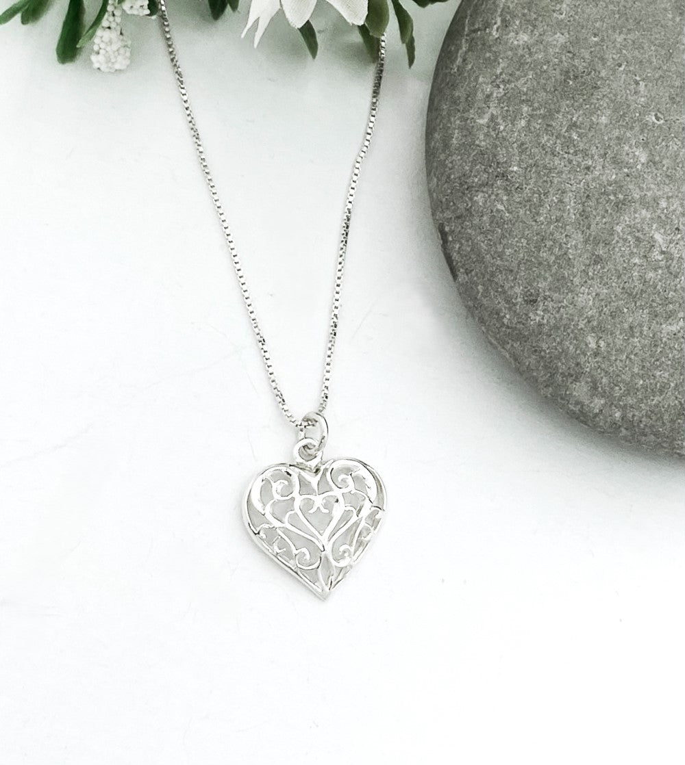 The Sterling Silver Filigree Heart Pendant, with an intricate design, is displayed next to a gray stone on a white background adorned with small white flowers. The 3/4 inch long pendant on a sterling chain pairs beautifully with matching earrings for an elegant set.