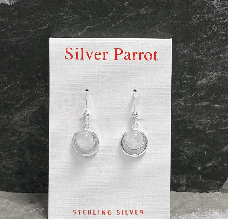 A pair of sterling silver earrings displayed on a card labeled "Silver Parrot." These exquisite Moonstone and Sterling Silver Delicate Dangles feature drop-style, round silver settings with translucent rainbow moonstone centers. The background is a textured dark green surface.