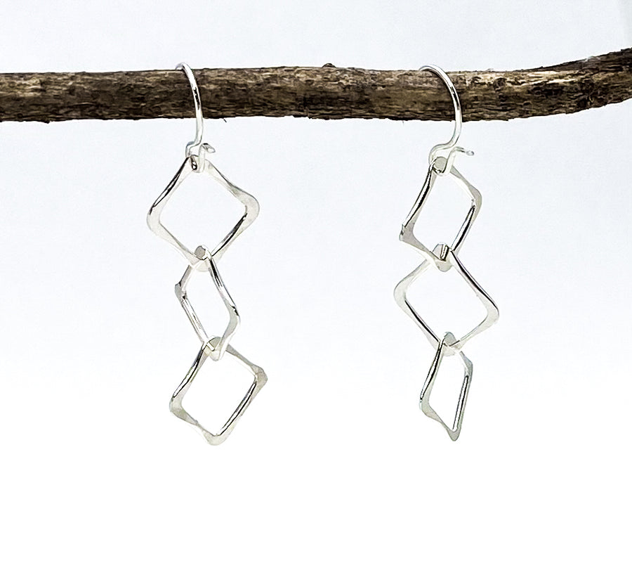 The "Sterling Silver 3 Hammered Squares" are handmade geometric dangle earrings from Kamas, UT. Featuring interconnected, angled square shapes hanging from a simple hook, these earrings measure 1.5 inches long and are displayed on a horizontal wooden branch against a plain white background.