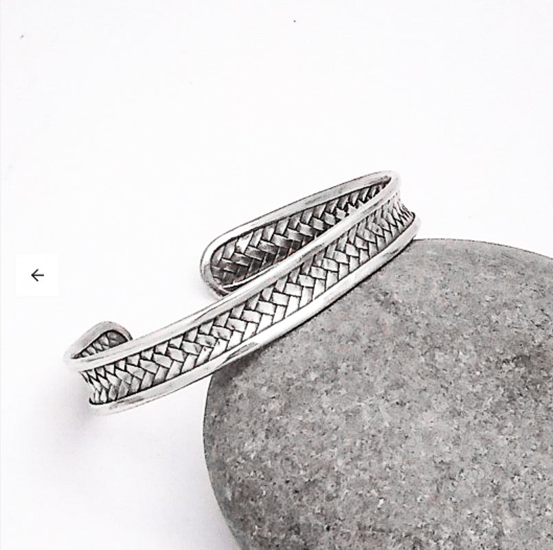 The Handmade Sterling Bali Woven Cuff, an intricately designed silver bracelet, rests gracefully on a smooth, gray stone. The cuff boasts a subtle and elegant texture that highlights its detailed craftsmanship. The plain white background accentuates the artistry of this hand-crafted accessory.