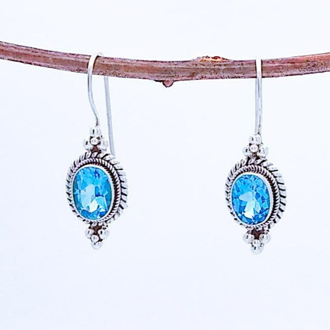 Oval Faceted blue topaz in sterling silver with a twist/braid detail around the edge and pyramids of 4 small silver balls on top and bottom edges. A wire extends from the top and can be closed in the back.