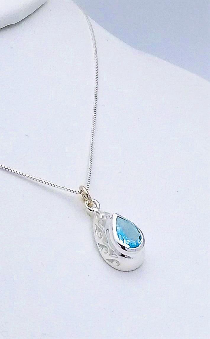 Sterling silver teardrop-shaped pendant with a simple design along the sides, and a blue topaz stone in the middle. Comes on an 18-inch chain
