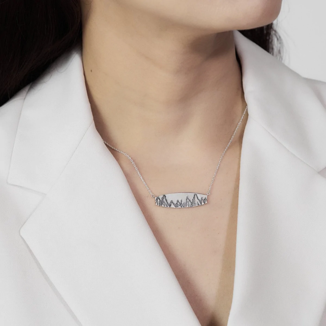 Flat rectangular sterling silver Boma necklace with engraving of mountains and trees etched along the bottom length. An adjustable rolo chain is attached to each side of the bar. Seen on woman with white blazer, silver necklace pops. 