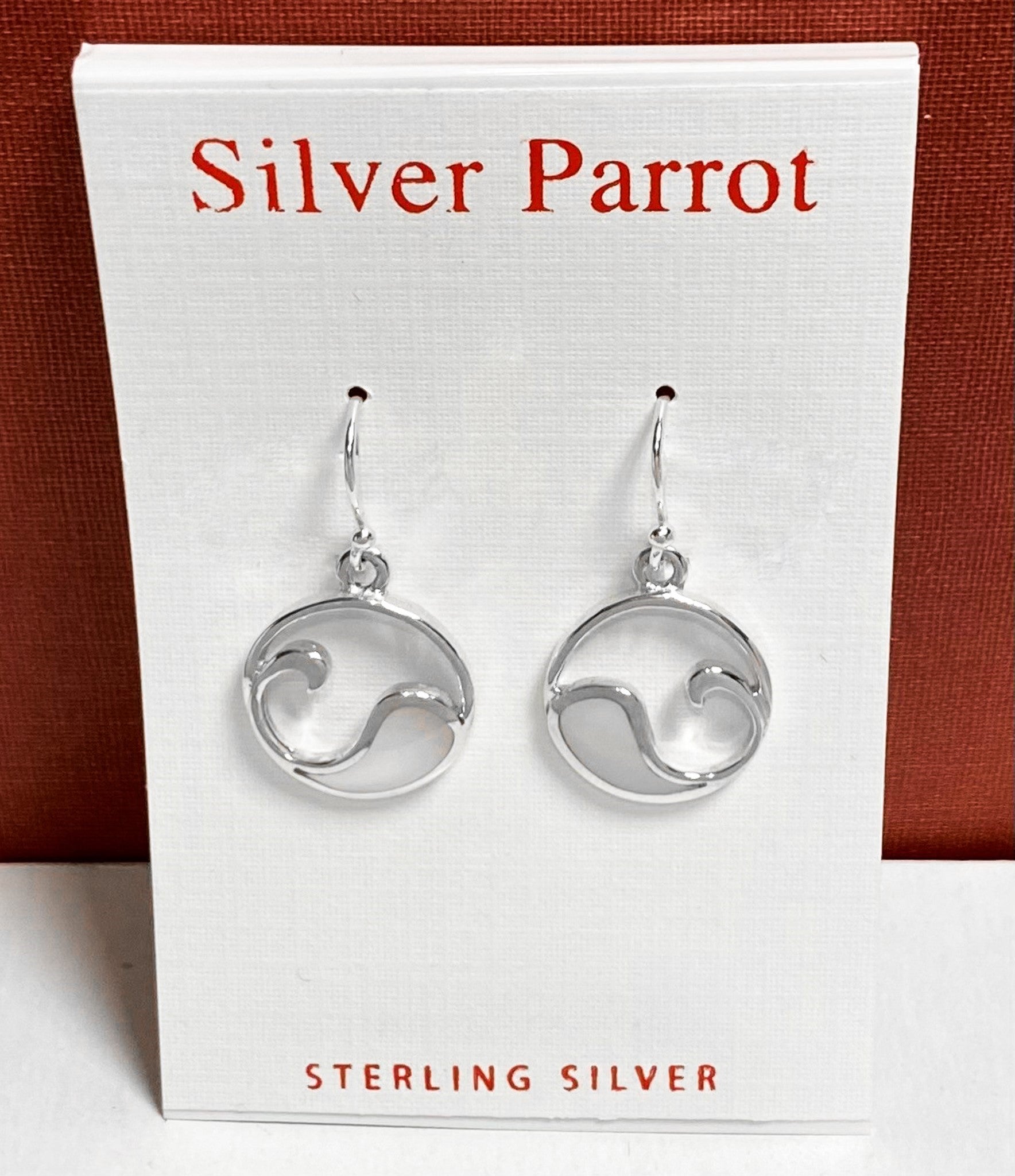 A pair of Great Wave Earrings with mother-of-pearl and sterling silver is displayed on a white card, featuring "Silver Parrot" written at the top in red text. These earrings boast a circular design with an abstract wave pattern reminiscent of the ocean. The display card is set against a red background.