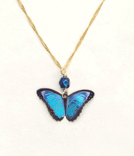 A bright blue realistic butterfly pendant with black outlines on a gold chain and a shiny dark blue bead.