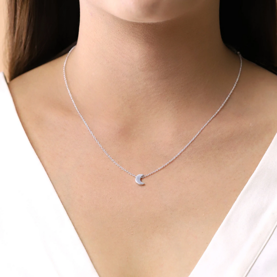 Small silver moon pendant on delicate silver chain, seen on human chest. Falls around the center of the collar bone.