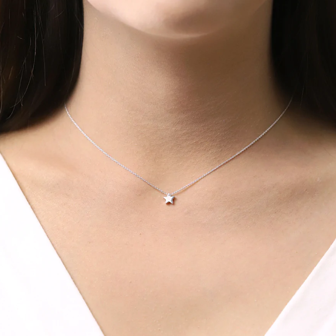 Small silver star pendant on delicate silver chain, seen on human chest. Falls around the center of the collar bone. 