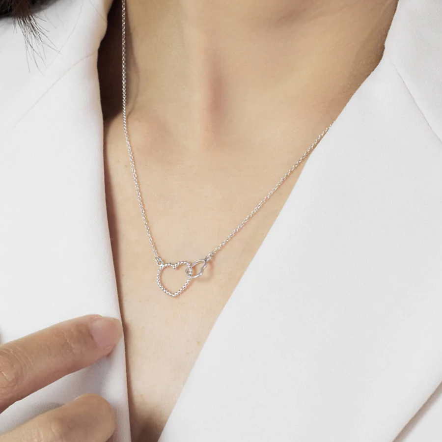 Interlocking heart necklace on woman's chest, one heart bigger than the other, bigger heart is textured while smaller heart is smooth; on a thin silver chain.