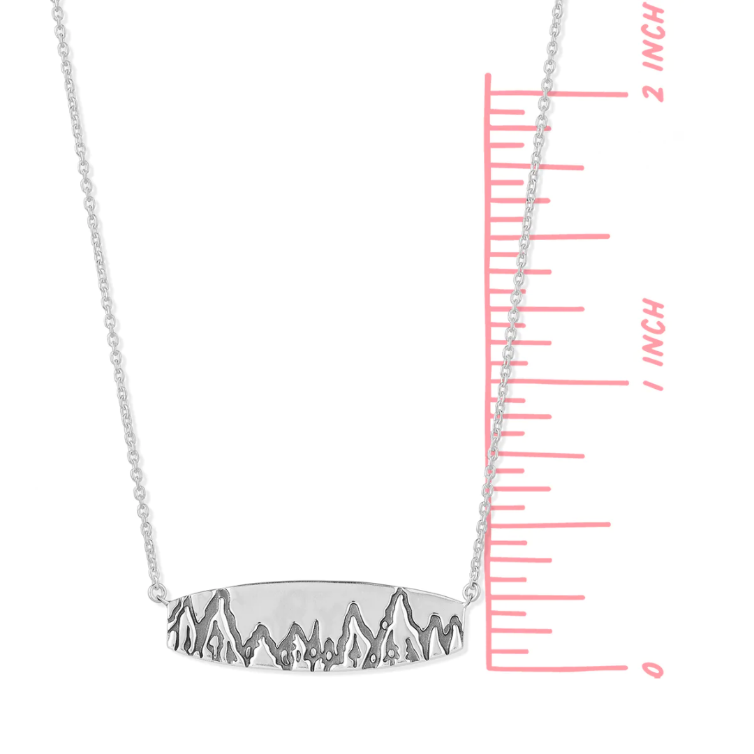 Flat rectangular sterling silver Boma necklace with engraving of mountains and trees etched along the bottom length. An adjustable rolo chain is attached to each side of the bar. Ruler is shown, seeing that pendant is one inch tall. 